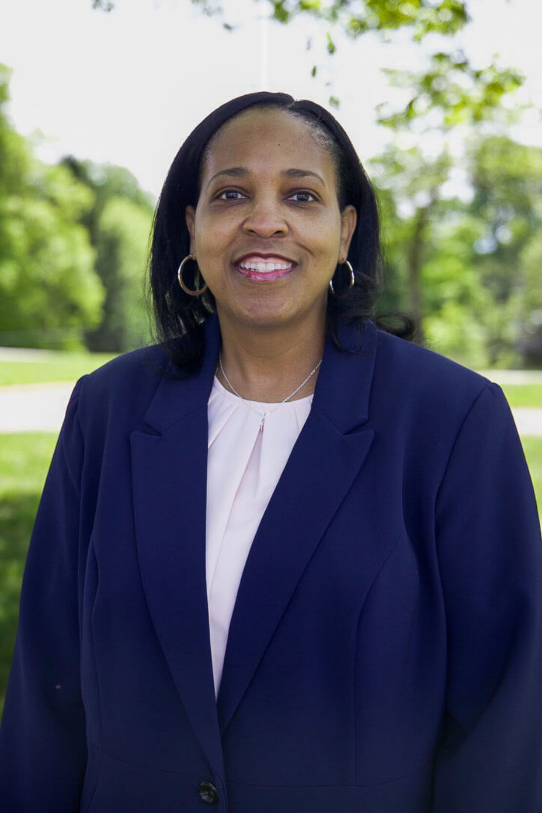 A woman wearing a navy blazer and light blouse smiles while standing outdoors, with green trees and grass in the background.