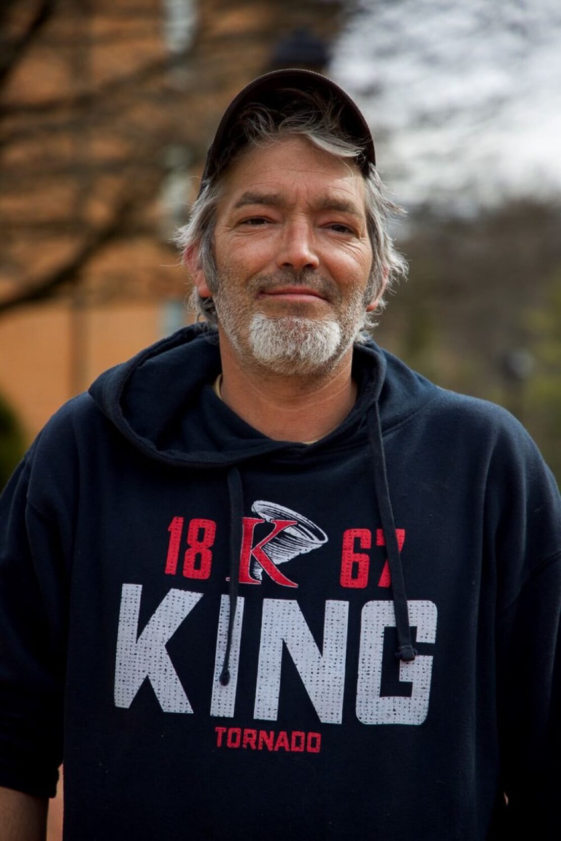 A man with gray hair and a beard wears a black hoodie with KING and Tornado printed on it, along with a black baseball cap. He is standing outdoors with a blurred background of trees and buildings.