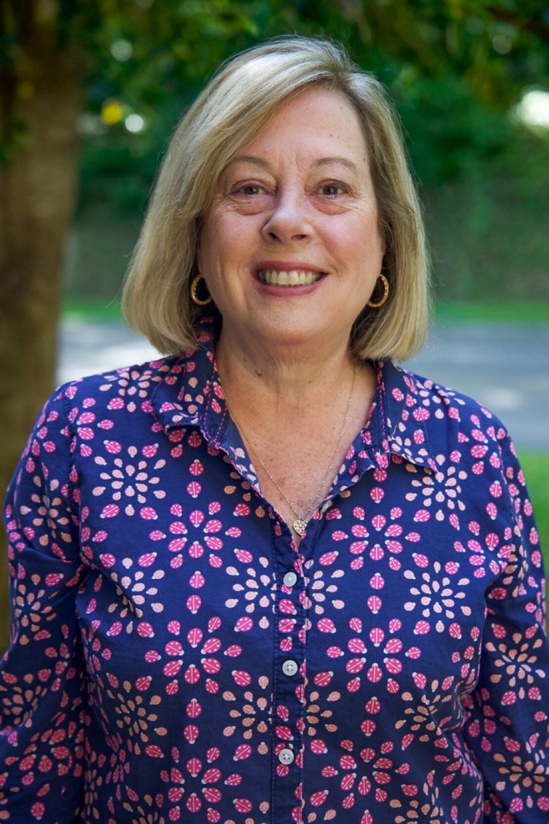 A smiling woman with shoulder-length blonde hair stands outdoors, wearing a blue shirt with a pink and white floral pattern. She is in front of greenery and a blurred background.
