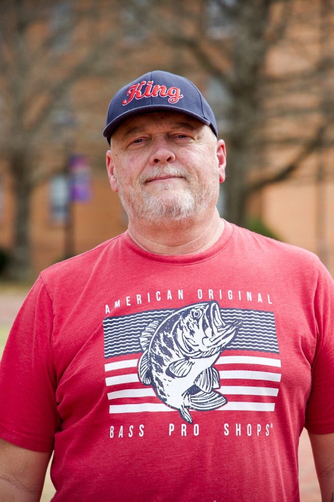 A man with a beard wears a navy King cap and a red Bass Pro Shops T-shirt featuring a large fish graphic, standing outdoors with blurred trees and buildings in the background.