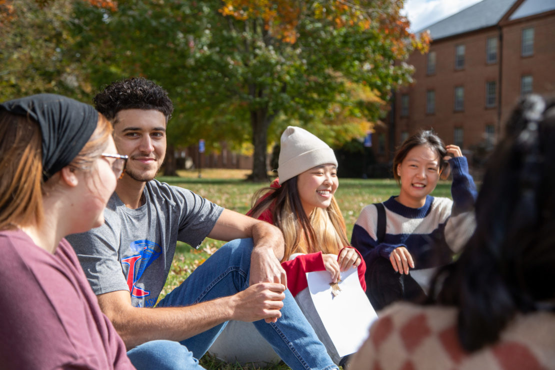 A group of college students sit in a circle on the grass, smiling and talking, with autumn trees and a brick building in the background.