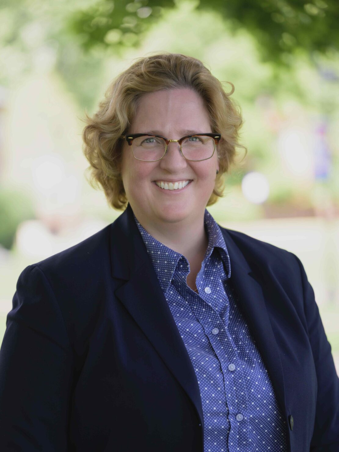 A person with short, curly blonde hair and glasses smiles outdoors. They are wearing a dark blazer over a blue patterned shirt, with greenery and trees blurred in the background.
