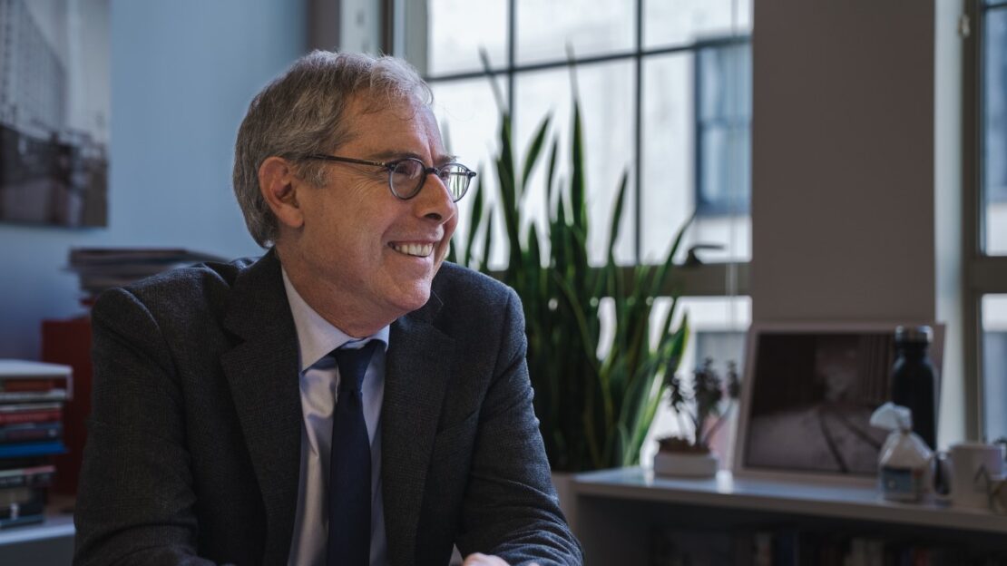 A man in a suit and glasses smiles while sitting at a desk in a well-lit office with large windows, books, a plant, and framed photos in the background.