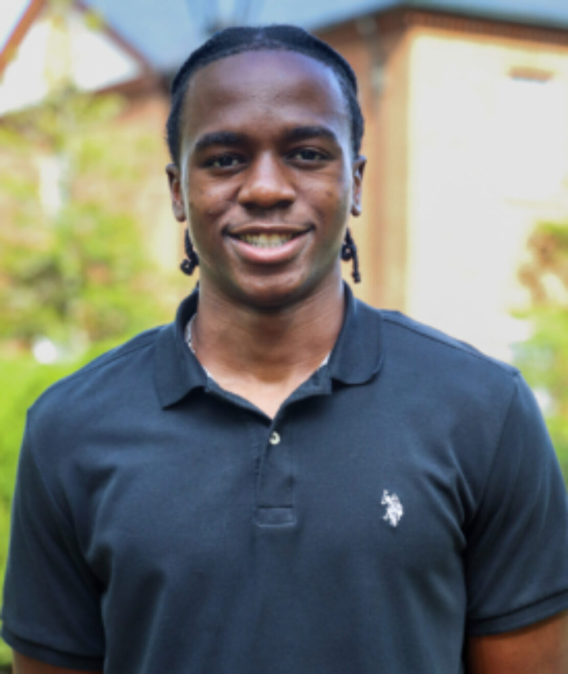 A young man with braided hair smiles while standing outdoors. He is wearing a black polo shirt and is positioned in front of blurred greenery and a brick building.