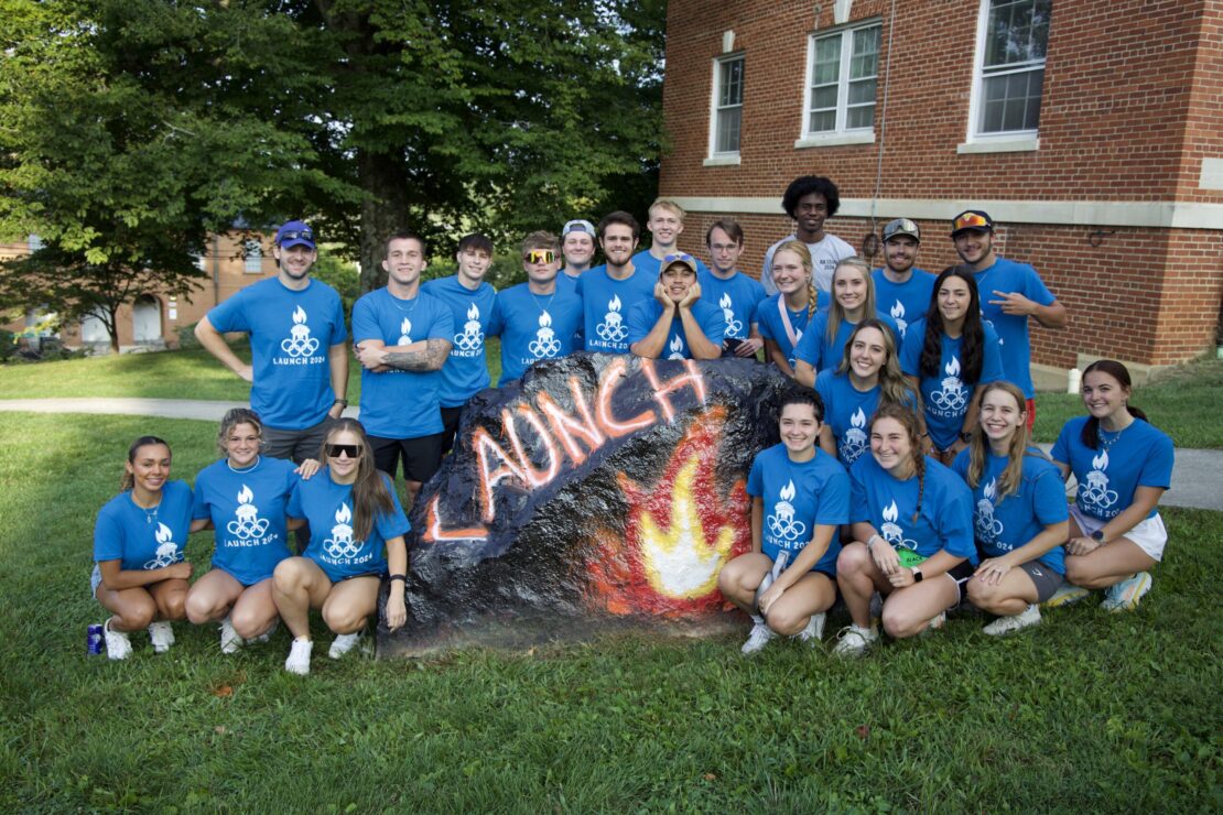 A group of smiling young adults in matching blue Launch T-shirts poses around a large rock painted with the word LAUNCH and a flame, in front of a brick building and trees on a sunny day.