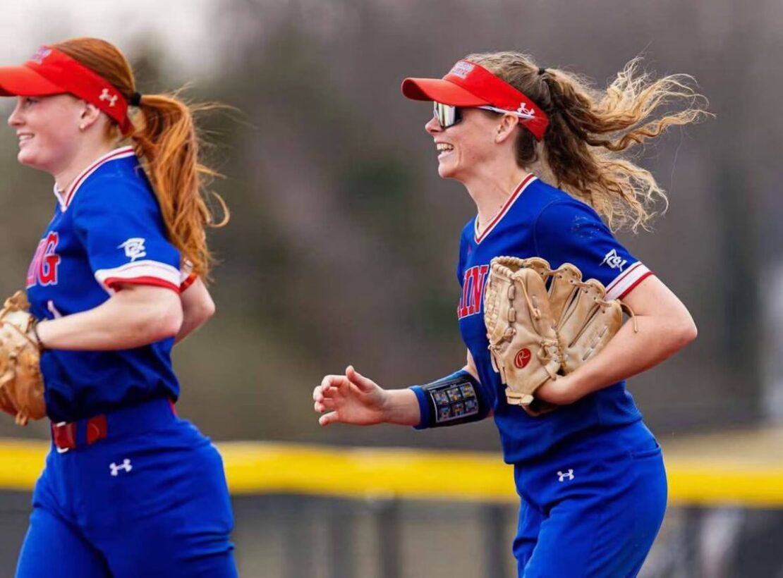 Two female softball players in blue uniforms and red visors run side by side, smiling, holding gloves. A yellow outfield fence and blurred trees are visible in the background.