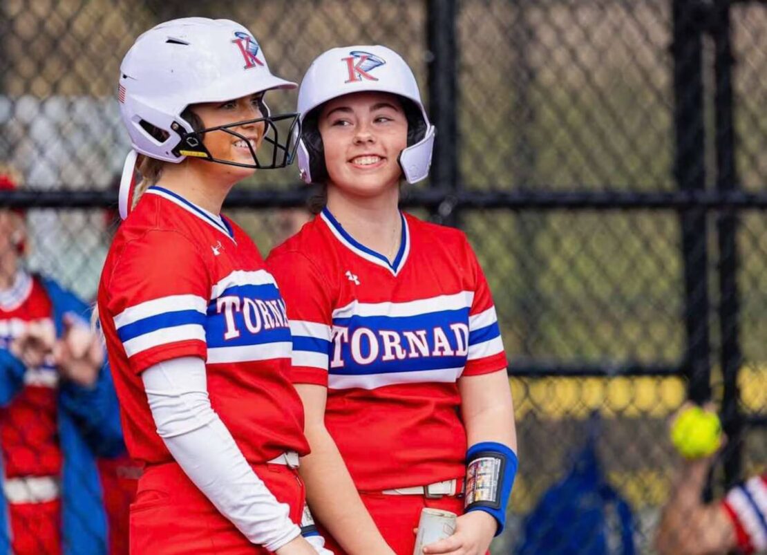 Two female softball players in red uniforms and white helmets stand and smile at each other near a chain-link fence, with TORNADO written on their jerseys.