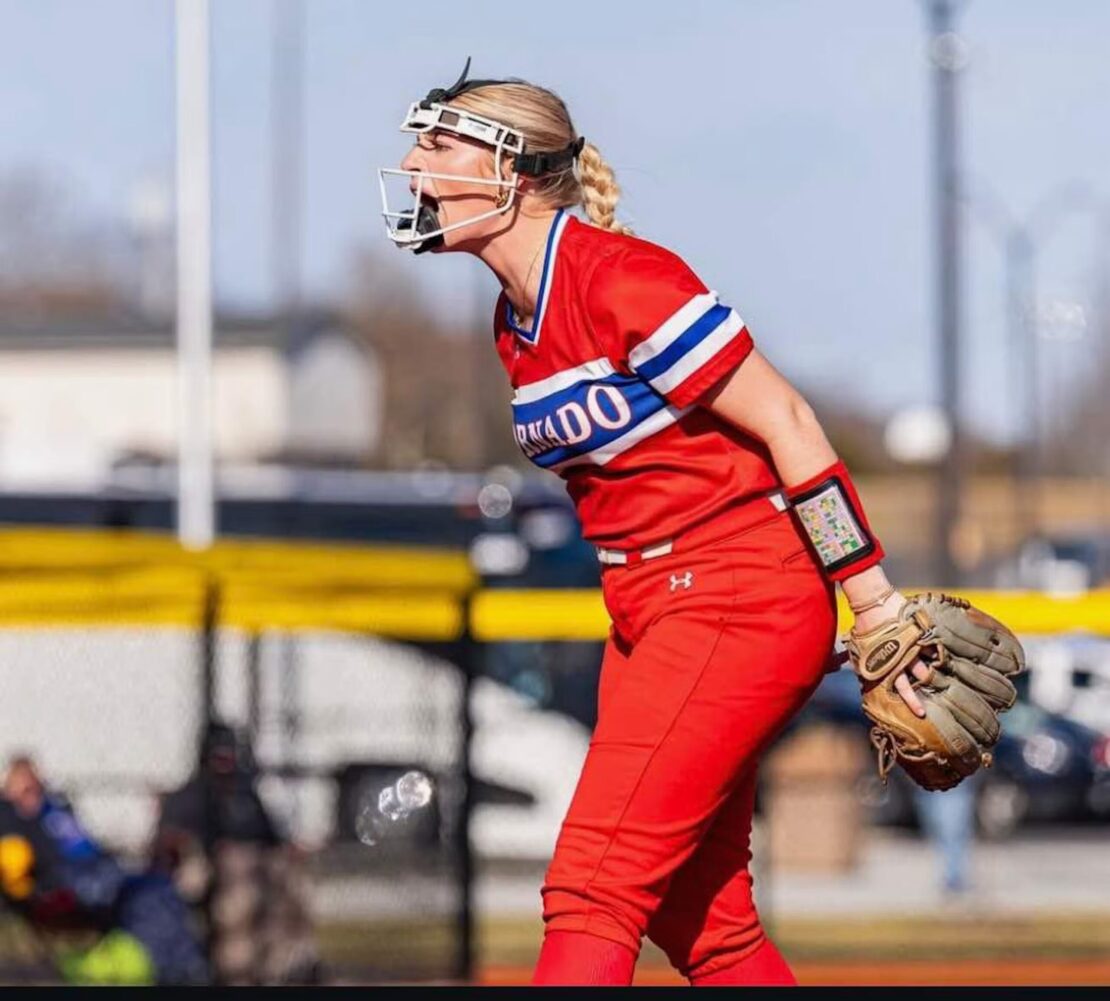 A softball player in a red uniform, wearing a face mask and glove, stands on the field and appears to be shouting or calling out, with a blurred background of a fence and spectators.