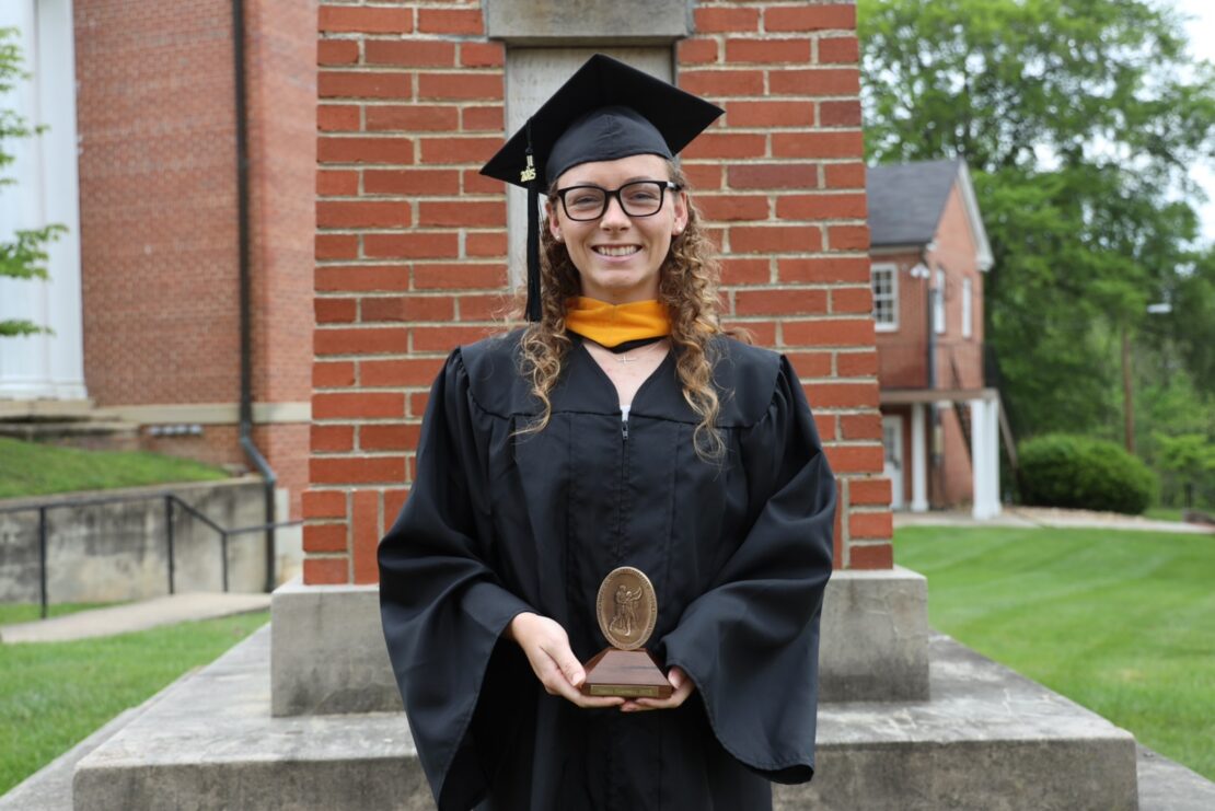 A graduate in a black cap and gown stands smiling, holding an award in front of a brick structure on a lawn with buildings and greenery in the background.