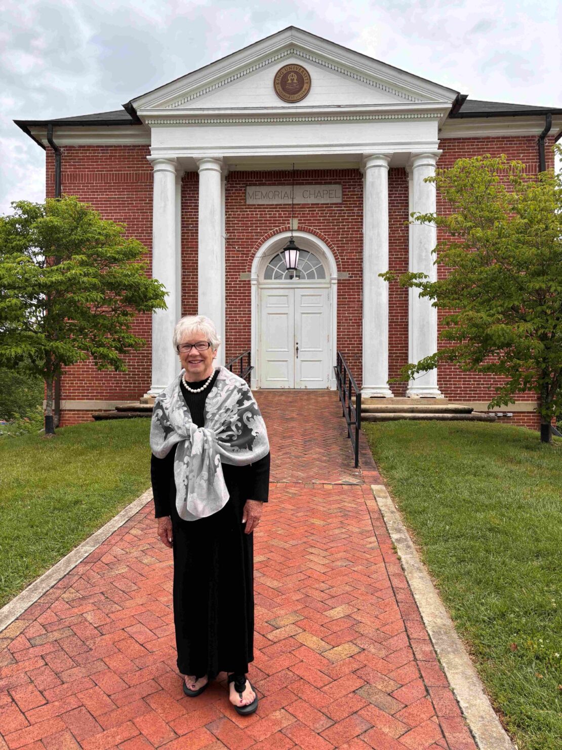 An older woman in a black dress and light shawl stands smiling on a brick walkway in front of a red-brick building with white columns labeled Memorial Chapel, surrounded by green trees.