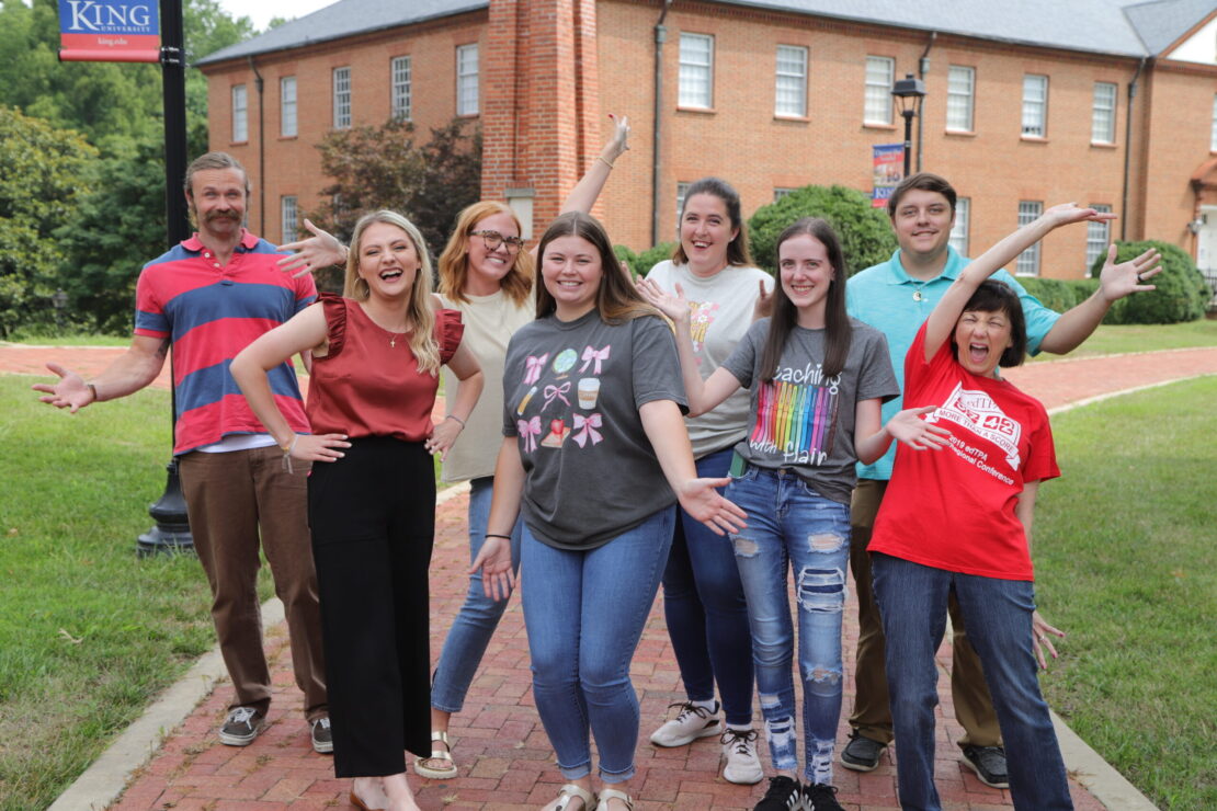 A group of eight smiling people pose enthusiastically on a brick pathway outside, in front of a building with red brick walls and several windows. Some have arms raised, and they all appear happy and excited.