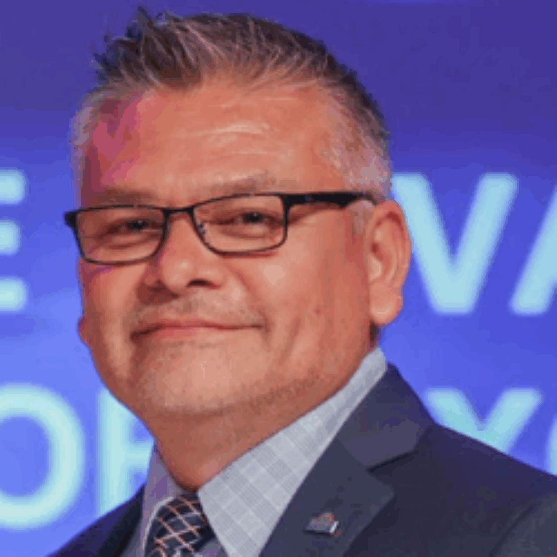 A middle-aged man with short gray hair, glasses, and a trimmed beard, wearing a blue suit, checkered shirt, and patterned tie, smiling in front of a blurred blue background with white text.