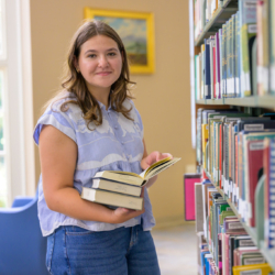 A young woman stands in a library, holding several books and reading one while smiling at the camera. Bookshelves and a window are visible in the background.