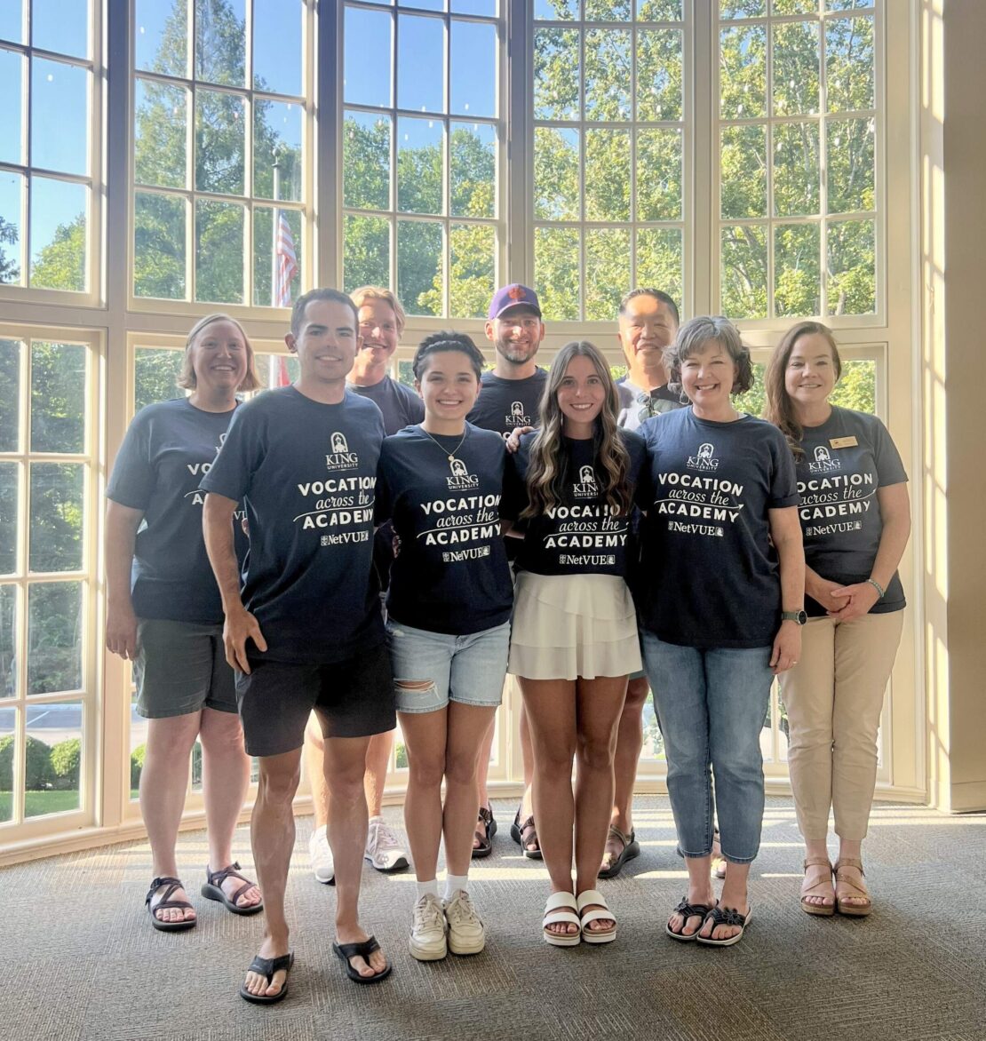 A group of ten people stands smiling in front of large windows with trees outside. Most wear matching dark blue Vocation Academy t-shirts, while one person in front wears a white shirt and skirt.