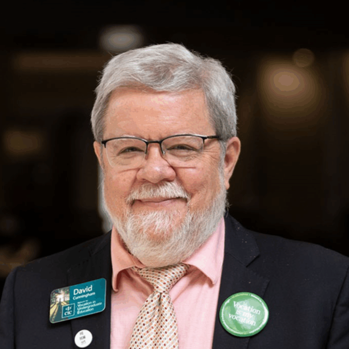 An older man with gray hair and a beard, wearing glasses, a black suit, pink shirt, and patterned tie. He has a David name tag and a green button that reads Vocation is vocation. He is smiling at the camera.