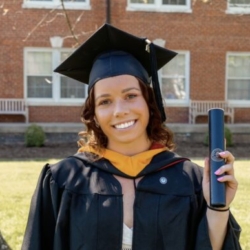 A young woman in a graduation cap and gown smiles while holding a diploma. She stands outside in front of a brick building with large windows and benches.