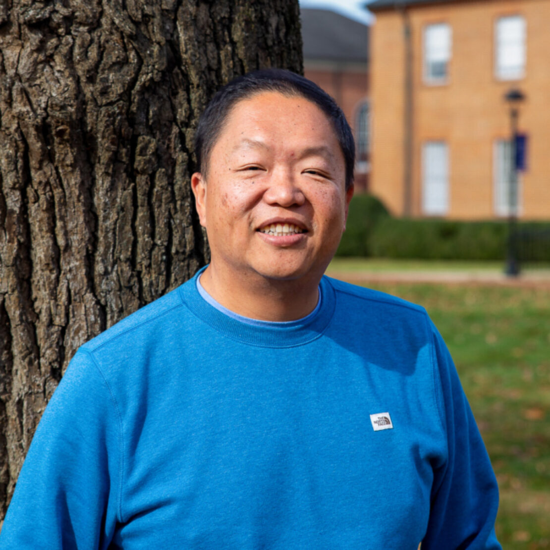 A person in a blue sweatshirt stands outdoors beside a tree, smiling at the camera. In the background, there is a brick building, green grass, and scattered autumn leaves.