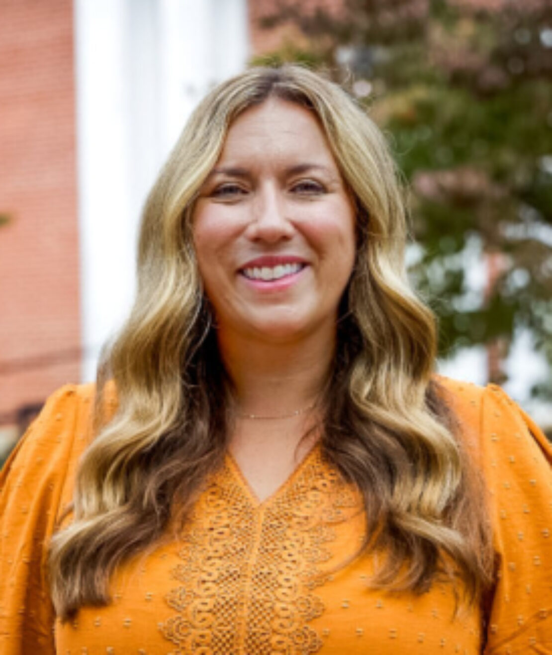 A woman with long, wavy blonde hair is smiling at the camera. She is wearing an orange blouse with intricate detailing, standing outdoors with blurred trees and a brick building in the background.