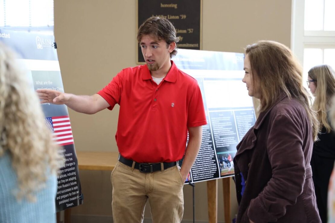 A man in a red polo shirt points to a presentation poster while speaking to a woman in a brown jacket. Other posters and people are visible in the background at what appears to be an indoor event.
