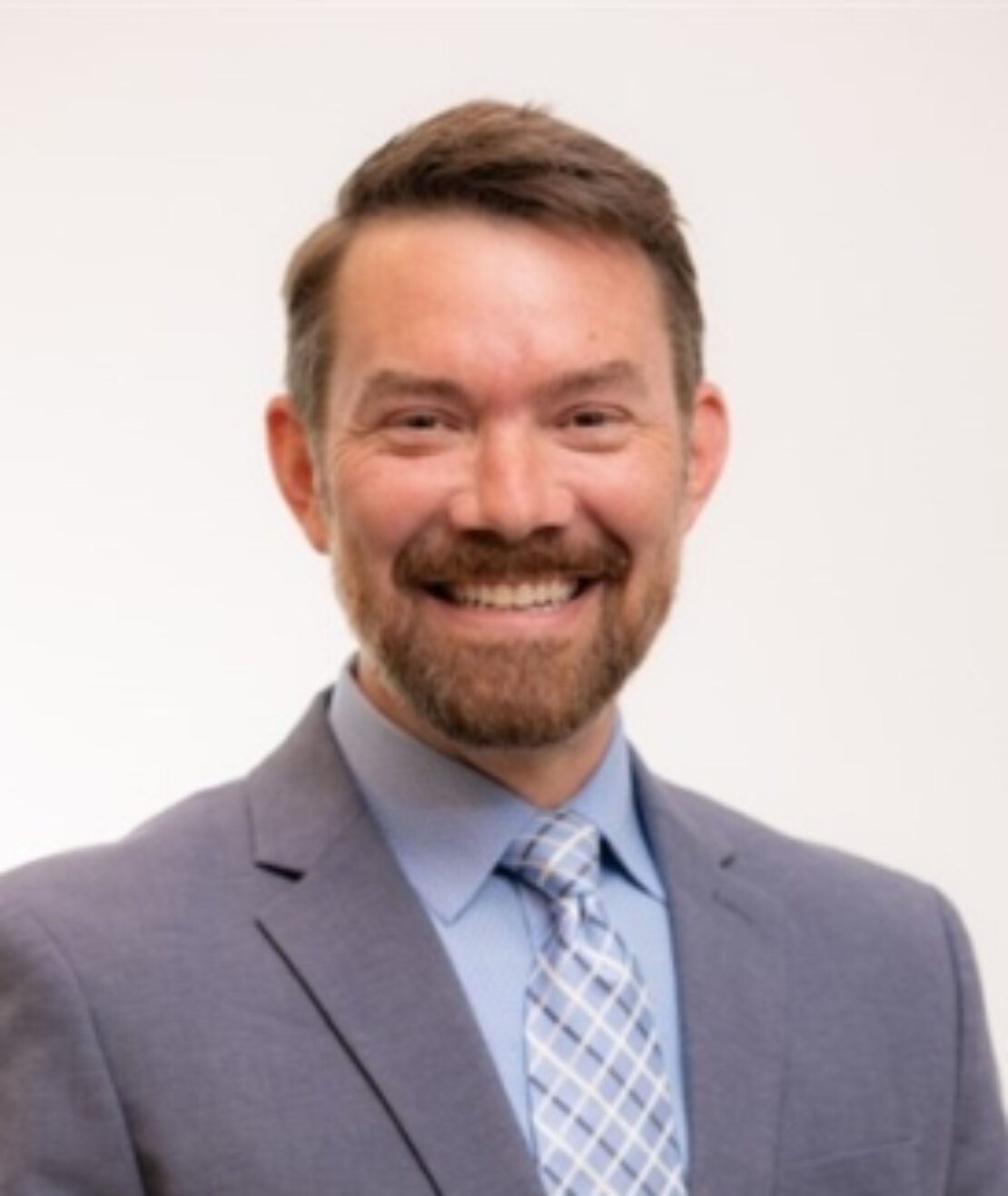 A man with short brown hair and a beard, smiling, wearing a light blue suit jacket, blue dress shirt, and a patterned blue tie, against a plain white background.