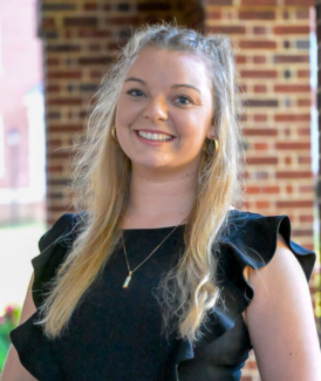 A woman with long blonde hair, wearing a black dress and gold necklace, stands smiling in front of a brick building with greenery in the background.