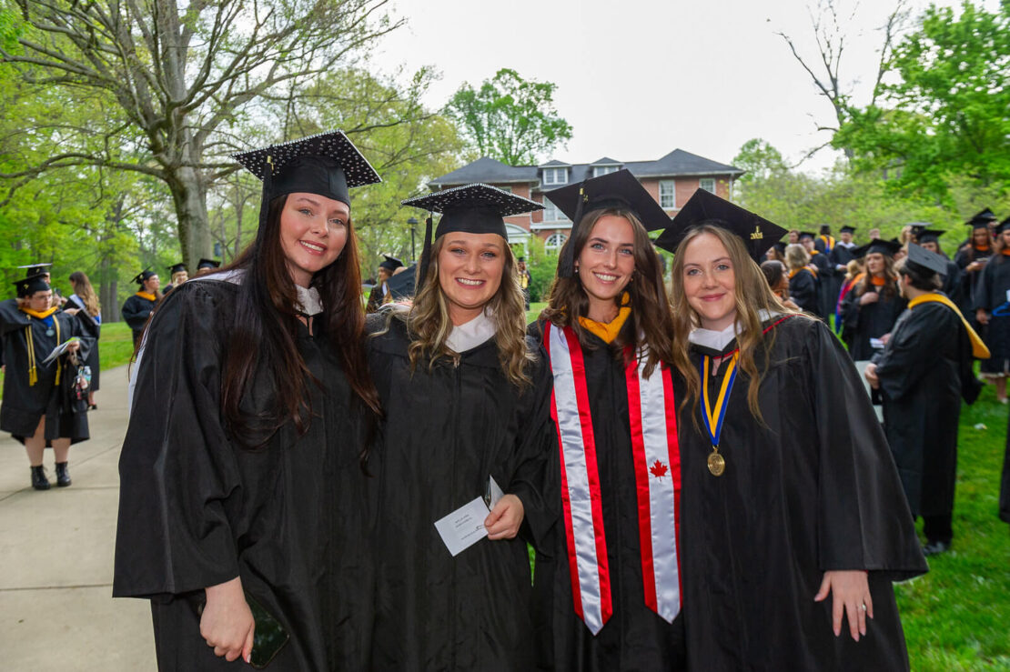 Four smiling graduates in caps and gowns pose outdoors, with trees and a crowd of other graduates in the background. One wears a white and red stole, and another has honor medals around her neck.