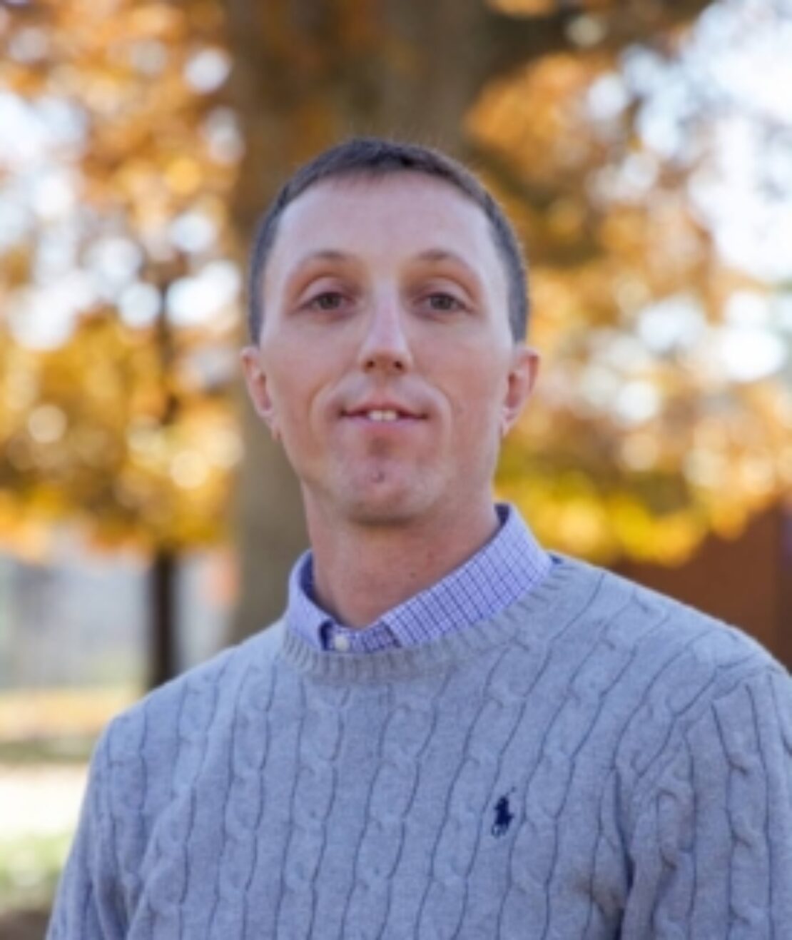 A man with short brown hair wears a light gray cable-knit sweater over a collared shirt, standing outdoors in front of trees with orange autumn leaves.
