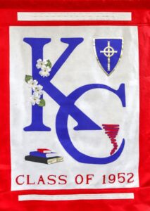 A banner with large blue letters KC, white flowers, a blue shield with a gold cross, three books, and the text CLASS OF 1952 in red on a white and red background.