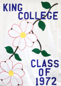 A white banner displays “KING COLLEGE CLASS OF 1972” in blue letters, with two large pink flowers and green leaves on a brown diagonal stem.