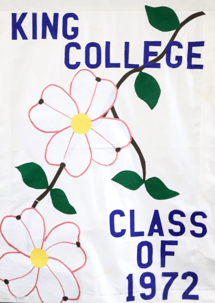 A white banner displays “KING COLLEGE CLASS OF 1972” in blue letters, with two large pink flowers and green leaves on a brown diagonal stem.
