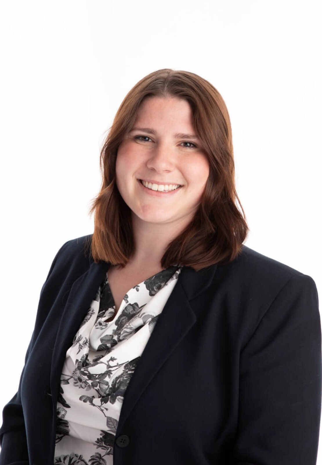 A woman with shoulder-length brown hair smiles at the camera. She is wearing a black blazer over a white blouse with a black floral pattern, posed against a plain white background.