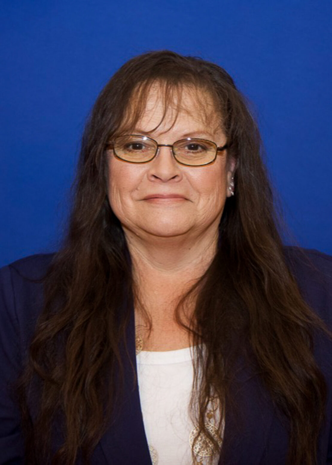 A woman with long brown hair and glasses, wearing a navy blazer over a white top, is standing in front of a solid blue background and smiling slightly.