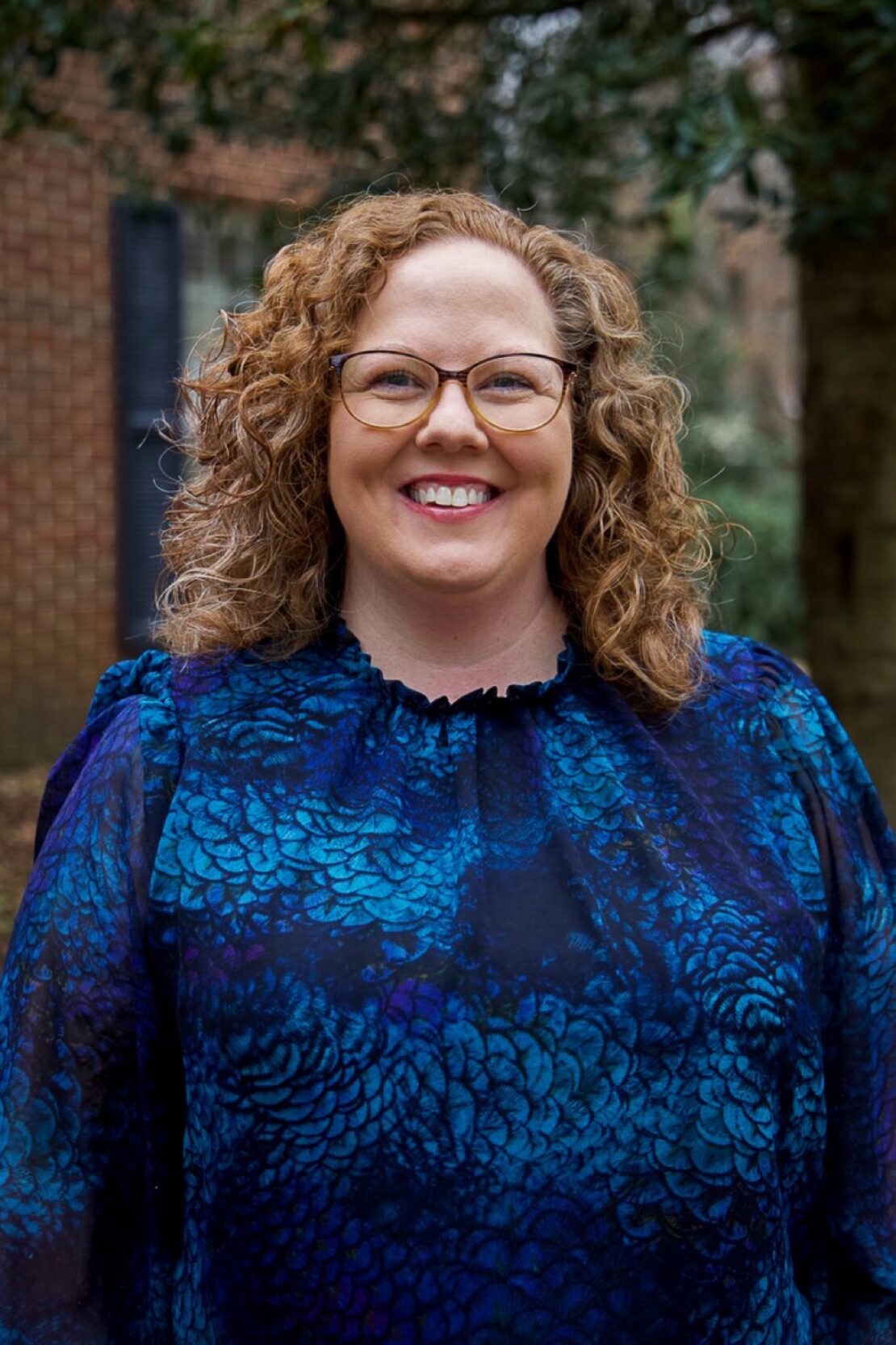 A woman with curly red hair and glasses smiles outdoors. She is wearing a blue patterned blouse, and trees and a brick wall are visible in the background.
