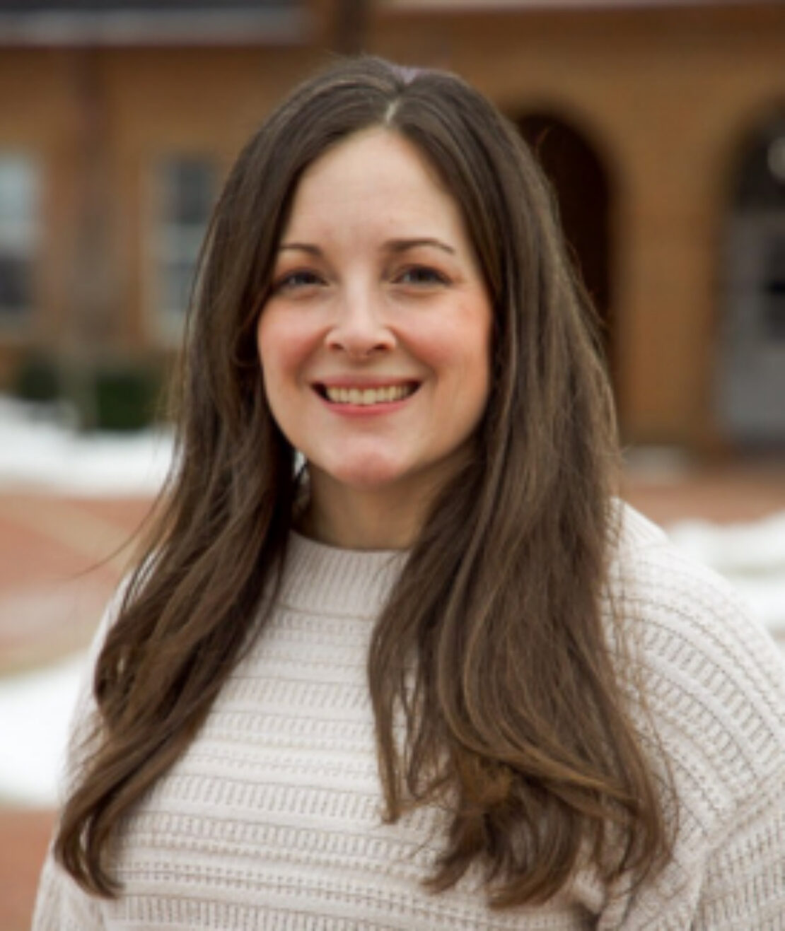 A woman with long brown hair wearing a light beige textured sweater stands outdoors, smiling at the camera. The background shows a brick building and patches of snow on the ground.