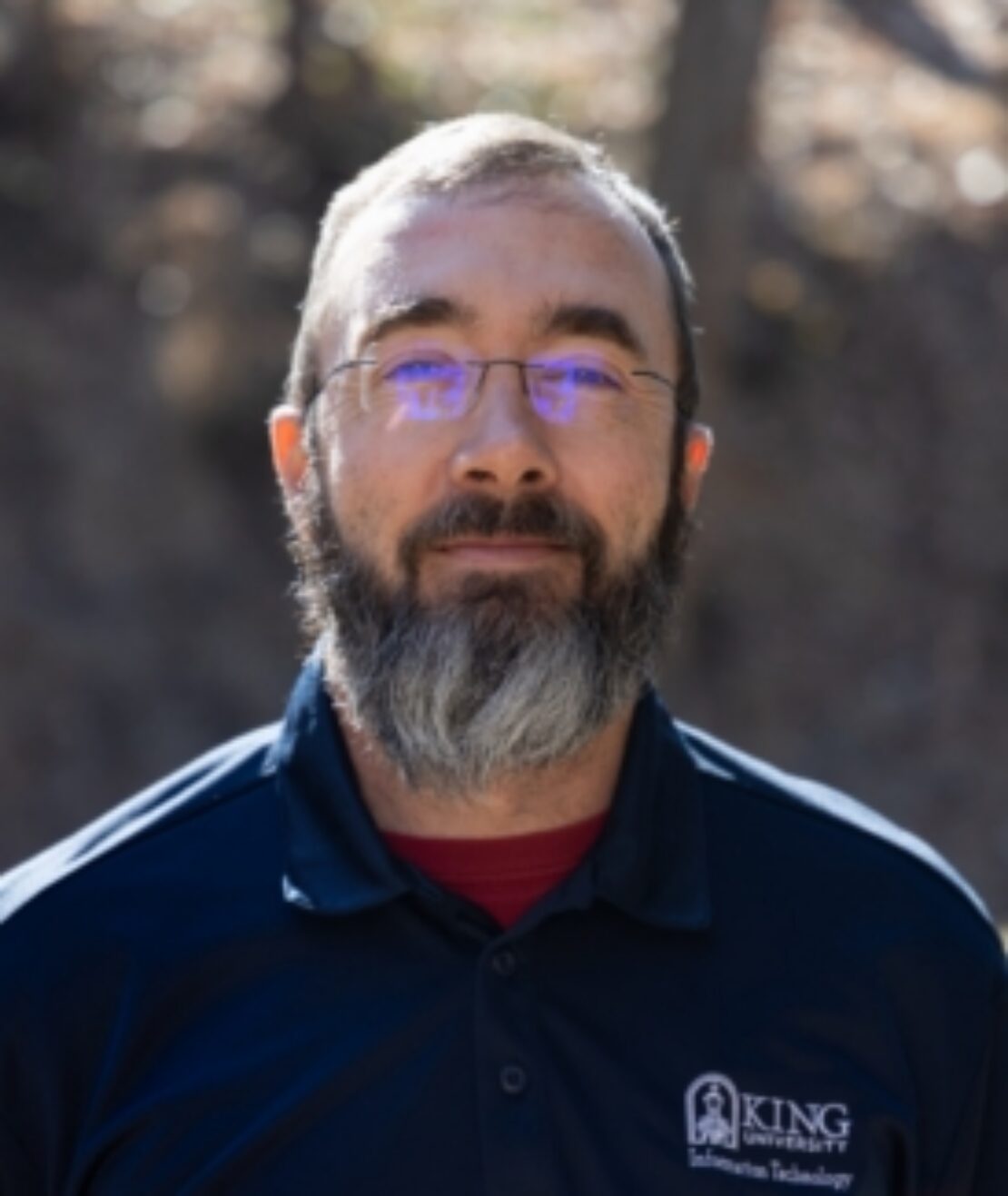 A bearded man wearing glasses and a navy blue King University Information Technology polo shirt stands outdoors with sunlight and trees in the background.