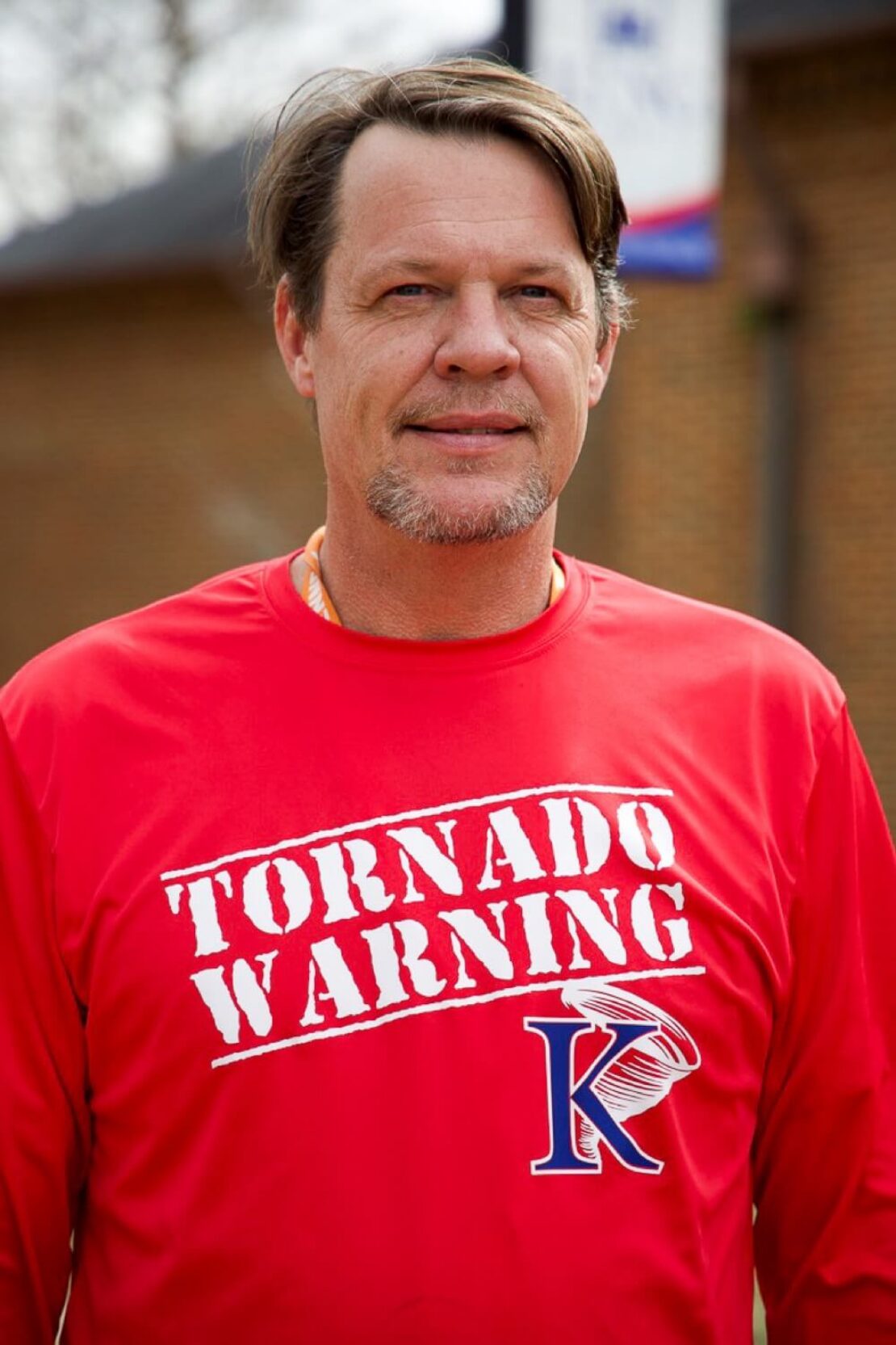 A man with short brown hair and a goatee wears a bright red shirt with TORNADO WARNING and a stylized K logo printed on the front, standing outside in front of a brick building.