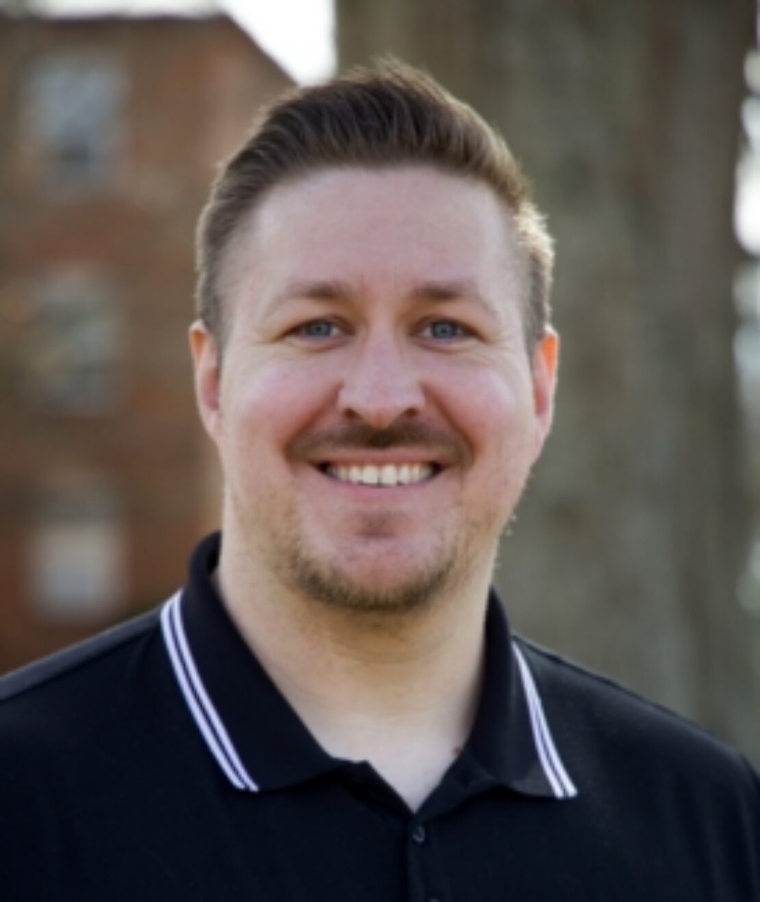 A smiling man with short light brown hair and a trimmed beard wears a black polo shirt with white stripes on the collar, standing outdoors with blurred trees and a building in the background.