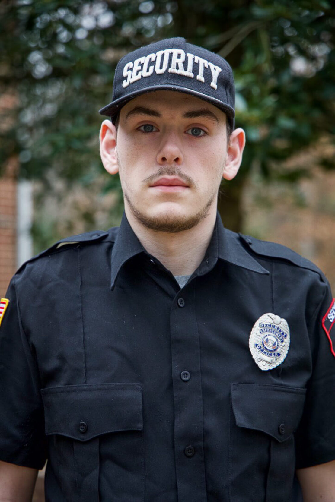 A young man wearing a black security uniform, cap with SECURITY written on it, and a badge, stands outdoors in front of blurred greenery and brick buildings.