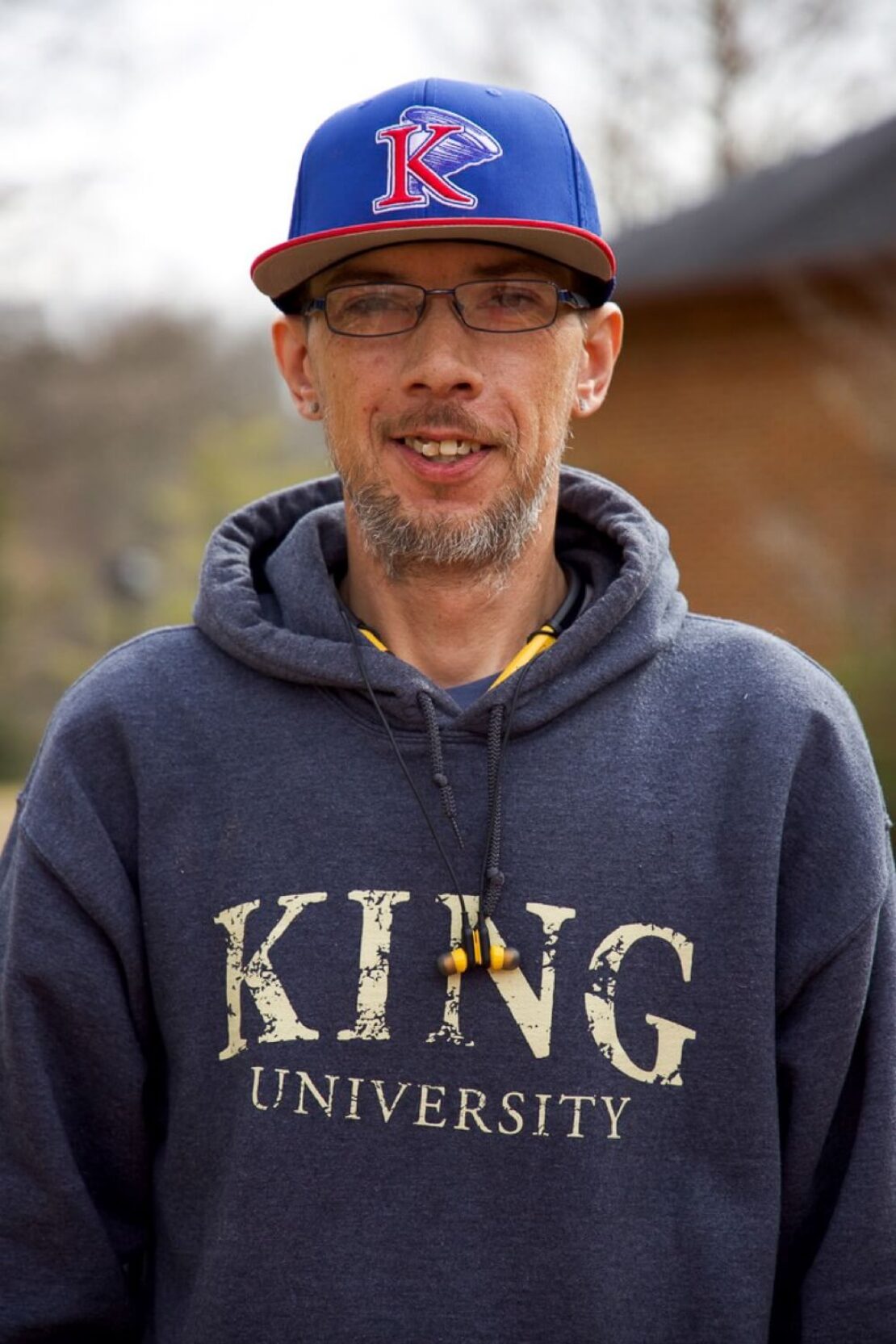 A man with glasses and a goatee wears a blue K cap and a dark King University hoodie, standing outdoors with a blurred background of trees and a brick building.