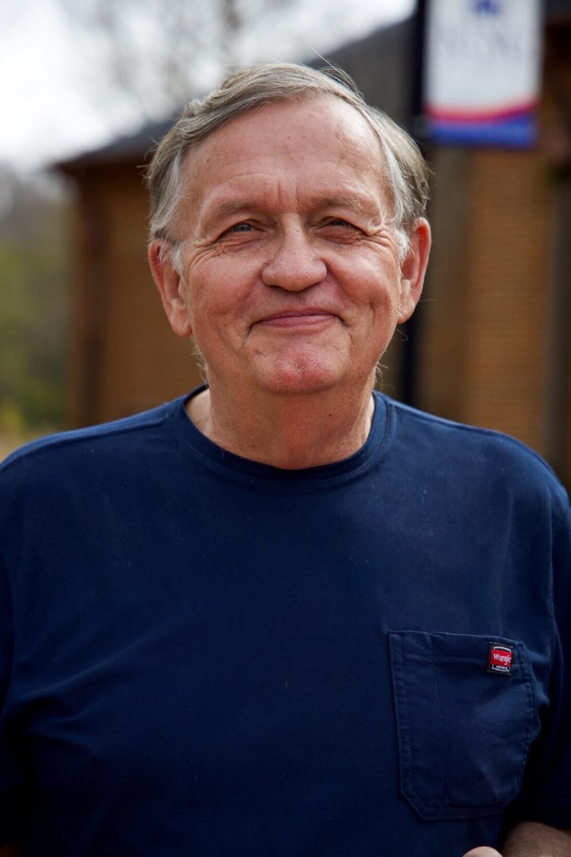 An older man with short gray hair is smiling gently outdoors. He is wearing a navy blue T-shirt with a small red logo on the pocket. The background is slightly blurred, showing a brick building and a banner.