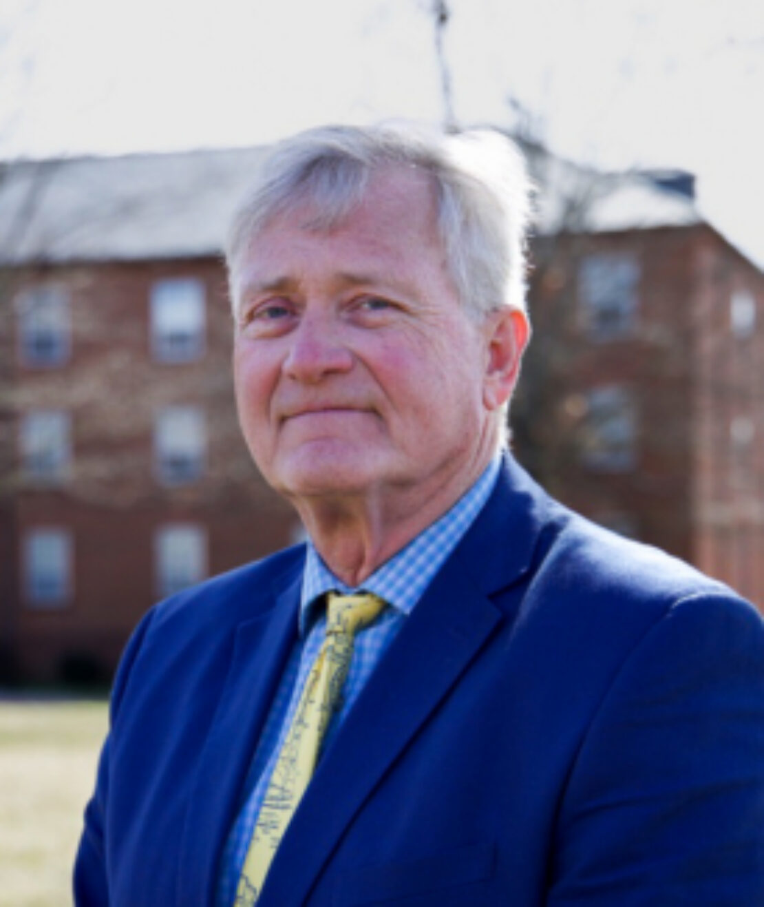 An older man with gray hair wearing a blue suit and yellow tie stands outside on a sunny day, with a brick building and bare trees in the background.