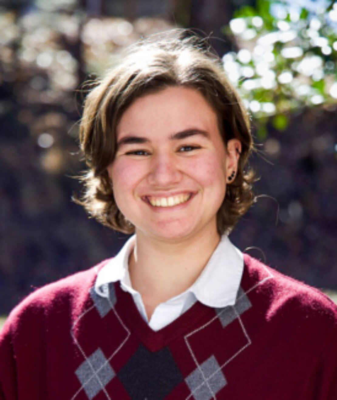 A person with short brown hair smiles outdoors, wearing a white collared shirt under a maroon sweater with a gray argyle pattern. Sunlight shines from behind, and the background is blurred with greenery.