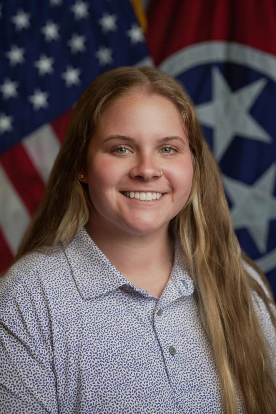 A smiling woman with long blond hair and a nose ring poses in front of American and Tennessee state flags, wearing a light patterned collared shirt.