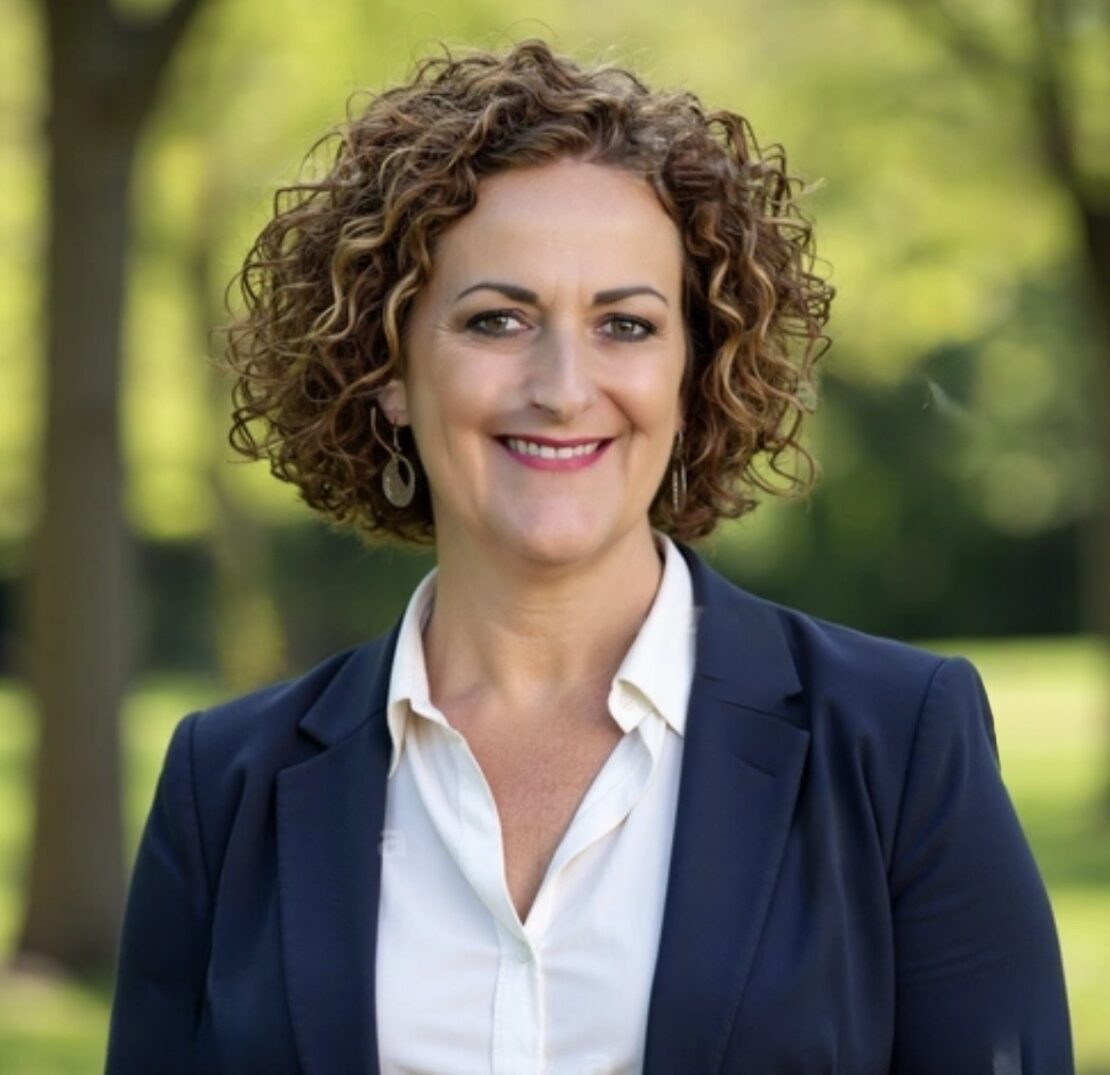 A woman with curly brown hair, wearing a navy blazer and white blouse, smiles while standing outdoors in a park with green trees in the background.