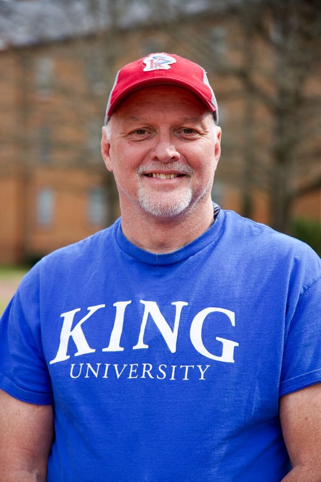 A smiling man wearing a red cap and a blue King University T-shirt stands outdoors, with a blurred building and trees in the background.