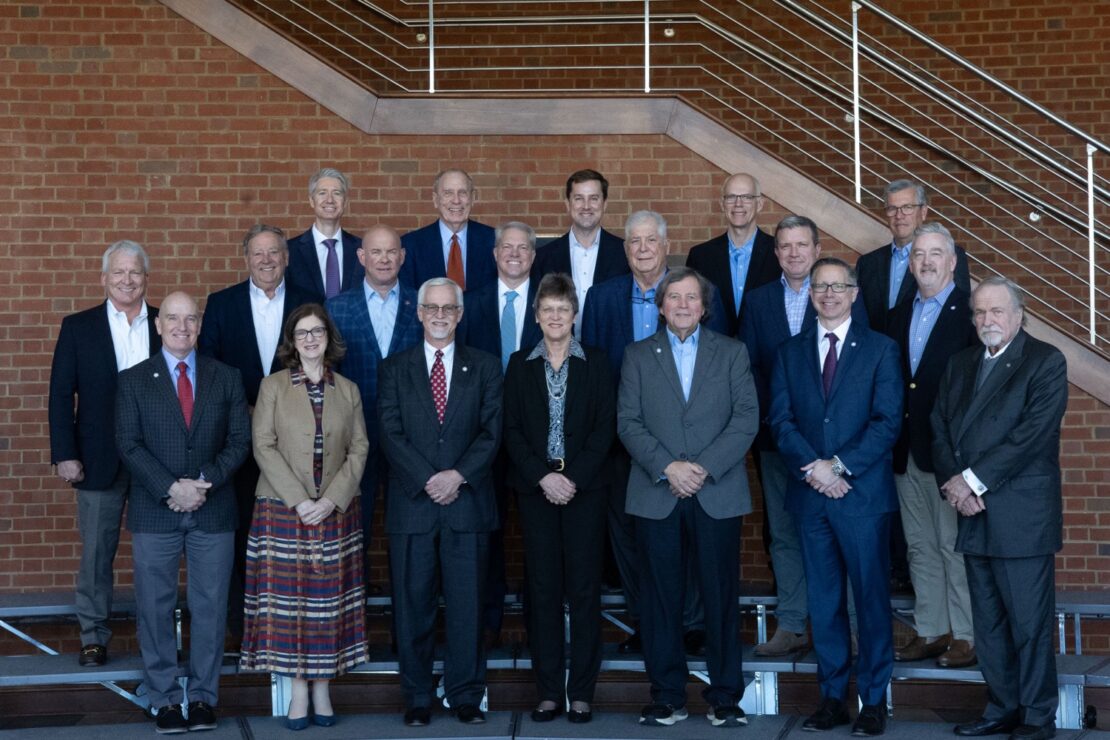 A group of 19 professionally dressed adults, including 3 women and 16 men, standing indoors in front of a brick wall and staircase, posing for a formal group photo.