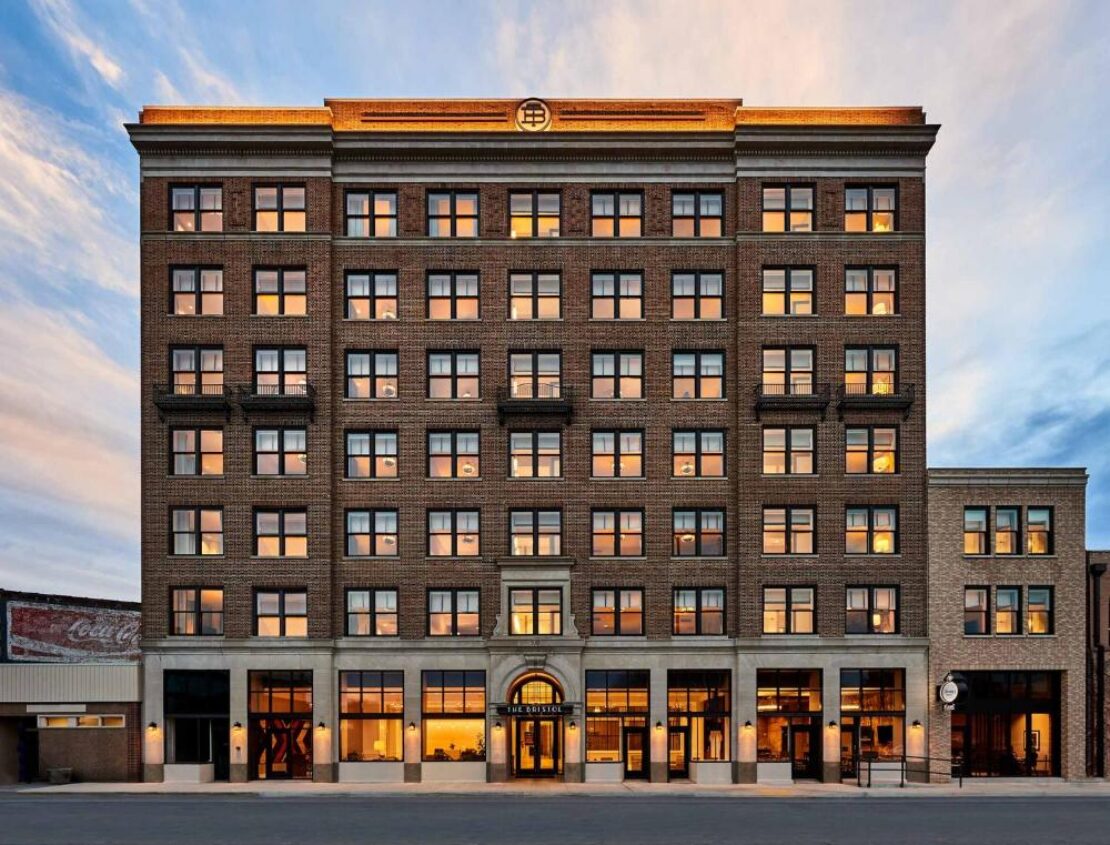 A six-story brick building with evenly spaced windows reflecting a sunset, featuring a central arched entrance and storefronts on the ground level, set against a partly cloudy sky.