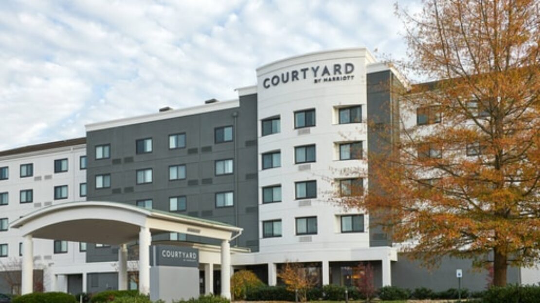 A modern Courtyard by Marriott hotel with four stories, gray and white exterior, large windows, an arched entrance, and a tree with orange leaves in the foreground.
