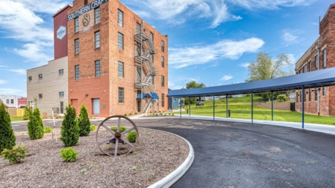 A red brick industrial building with an exterior metal staircase stands beside a landscaped area featuring small shrubs and a large wagon wheel, with a covered driveway curving around the scene under a blue sky.