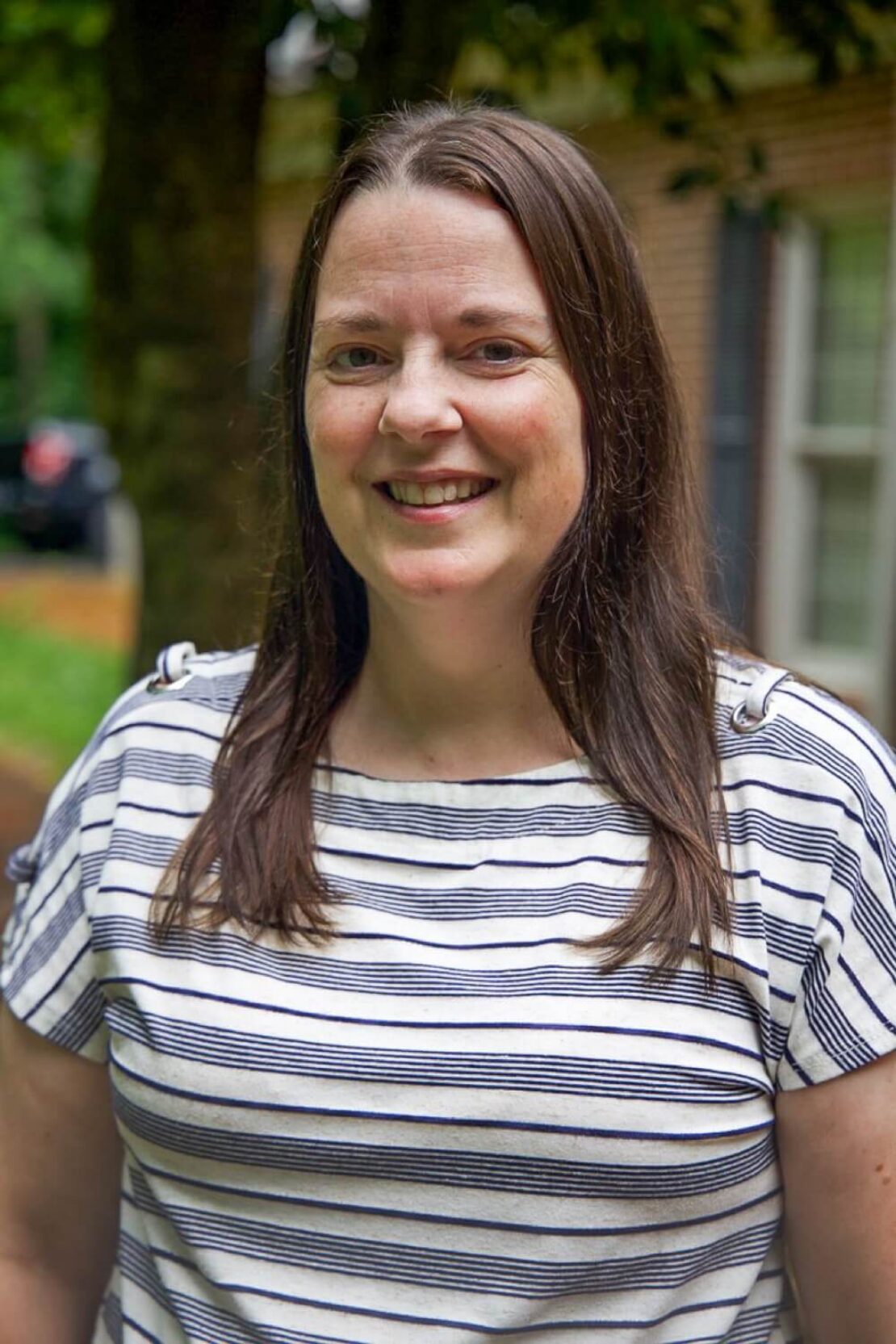 A woman with long brown hair, wearing a white and navy striped shirt, smiles outdoors. There are trees and a building with windows in the blurred background.